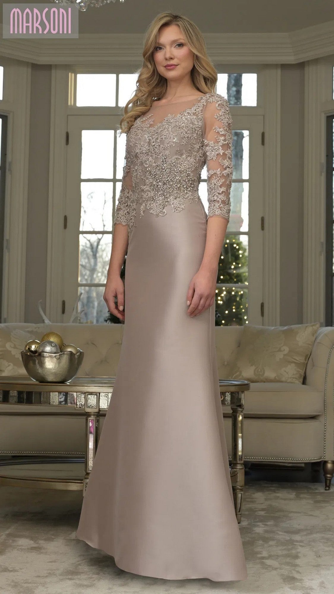 A woman poses gracefully indoors in the Marsoni MV1357, a long taupe formal dress featuring lace-embroidered bodice and sleeves, standing before a sofa and large windows.